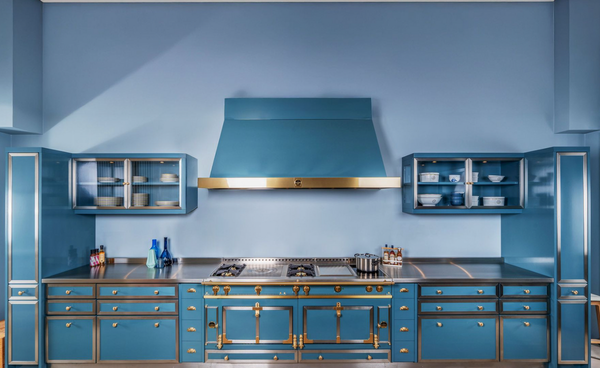 A luxury kitchen cabinetry set in a rich teal blue with ornate gold hardware, featuring a central range hood, gas stove, and matching side cabinets against a blue wall.