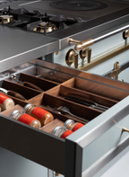 Close-up of a stainless steel kitchen range with an open drawer containing wooden utensil organizers and spice jars with red lids.