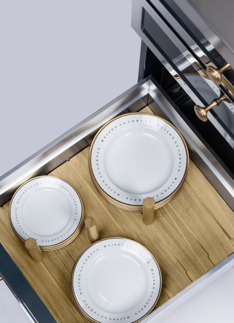 Three white ceramic plates with gold-rimmed edges displayed on a wooden stand inside a stainless steel drawer or cabinet.