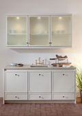 A white kitchen vanity cabinet with three upper glass-front cabinets and three lower drawers, styled with a marble countertop, brass faucet, and decorative items, set against a warm beige wall.