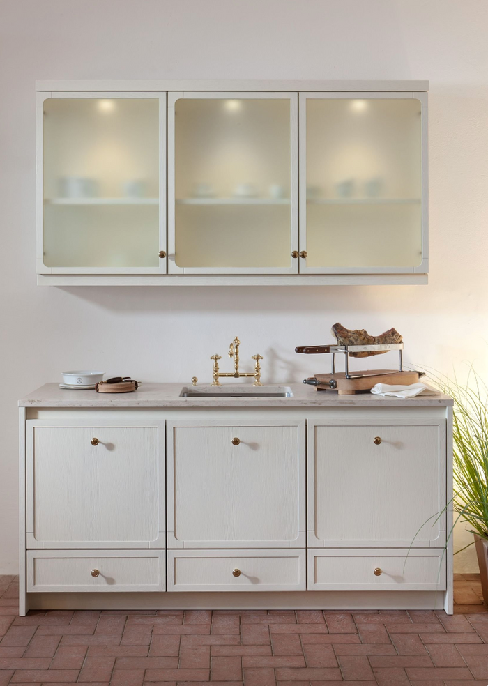 A white kitchen vanity cabinet with three upper glass-front cabinets and three lower drawers, styled with a marble countertop, brass faucet, and decorative items, set against a warm beige wall.