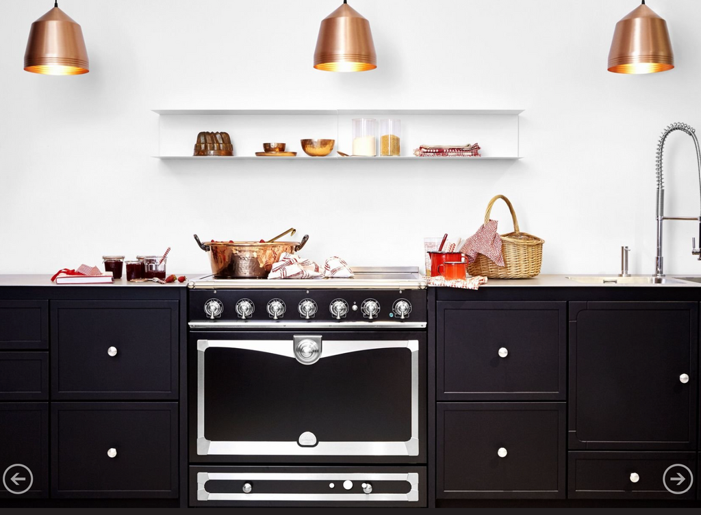A black freestanding gas range with a large oven door and silver control knobs, set in a modern kitchen with dark cabinets, a white wall, copper pendant lights, and countertop cookware.