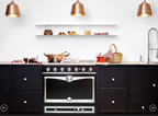 A black freestanding gas range with a large oven door and silver control knobs, set in a modern kitchen with dark cabinets, a white wall, copper pendant lights, and countertop cookware.