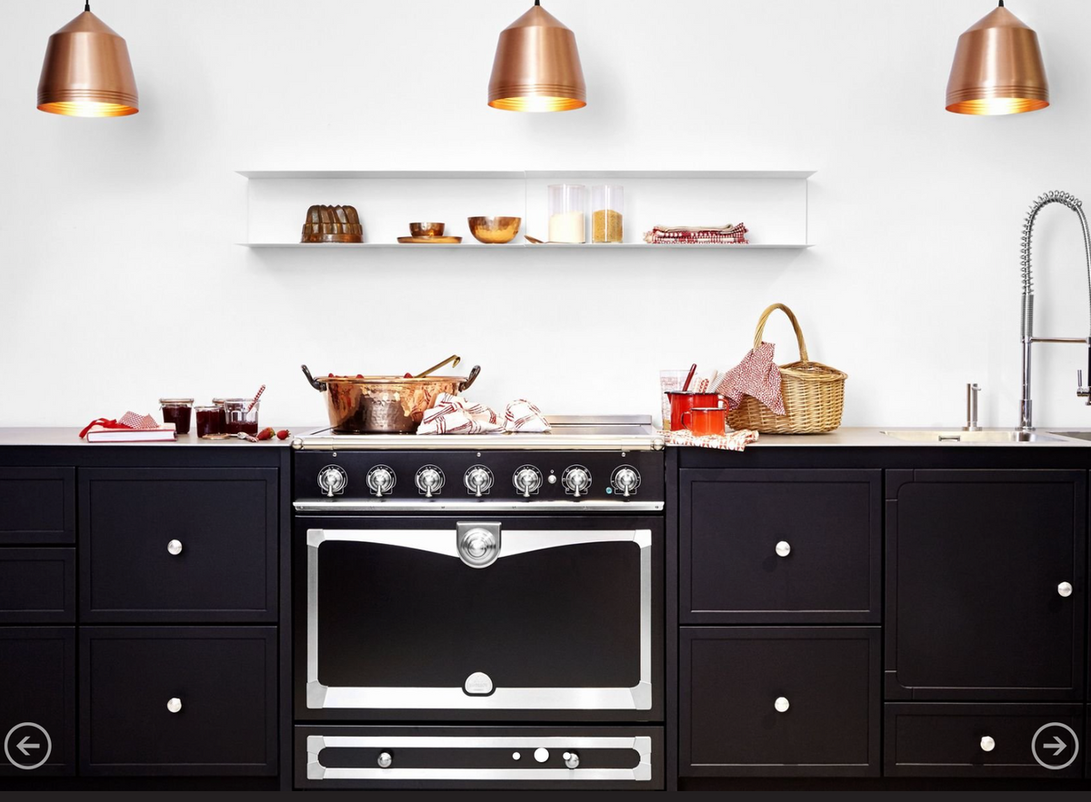 A black freestanding gas range with a large oven door and silver control knobs, set in a modern kitchen with dark cabinets, a white wall, copper pendant lights, and countertop cookware.