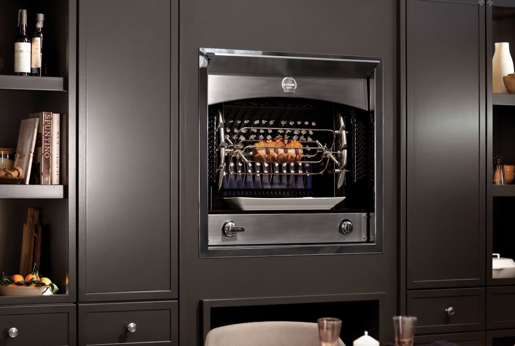 A built-in stainless steel gas grill with a rotisserie rod and tray, set into a dark charcoal kitchen cabinet with dining table props visible in the foreground.