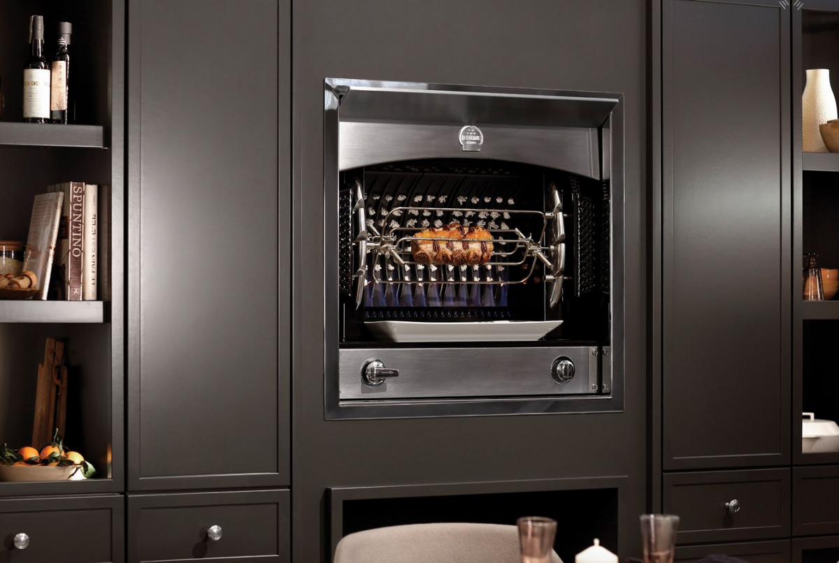 A built-in stainless steel gas grill with a rotisserie rod and tray, set into a dark charcoal kitchen cabinet with dining table props visible in the foreground.