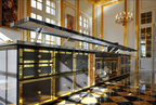 A long, luxurious kitchen island or prep station with black surfaces and extensive gold trim, set in an ornate room with gilded walls, checkered marble flooring, and chandeliers.