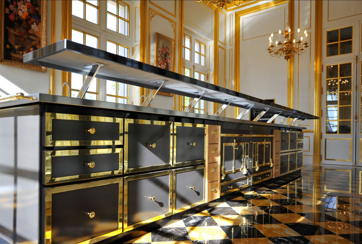 A long, luxurious kitchen island or prep station with black surfaces and extensive gold trim, set in an ornate room with gilded walls, checkered marble flooring, and chandeliers.